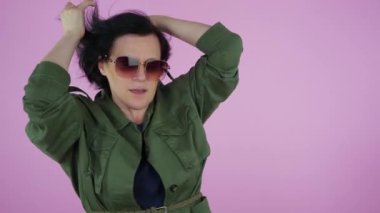 Yeşil elbiseli bir kadın, gözlük takıyor, dans ediyor, elleri müziğin ritmine göre hareket ediyor, rüzgar saçlarını savuruyor. Pembe arka plan. Yavaş çekim.
