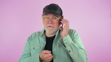 Beyzbol şapkalı, gözlüklü, mavi tişörtlü, telefonda konuşan, duygusal olarak hareket eden ve gülümseyen yaşlı bir adam. Pembe arka plan. Yavaş çekim.