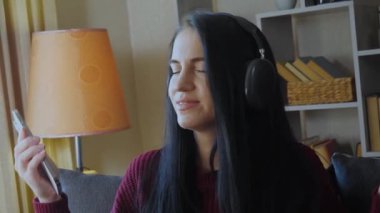 Koyu renk saçlı, bordo kazaklı, kulaklıklı, müzik dinleyen, telefon kullanan, duygusal olarak ritme göre sallanan ve gülümseyen güzel bir kız. Ev ortamı.