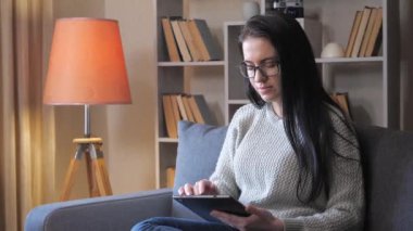 Genç, güzel bir kadın, evinde kanepede oturuyor, tablet üzerinde iletişim kuruyor, duygusal olarak hareket ediyor. Sıcak ev ortamı.