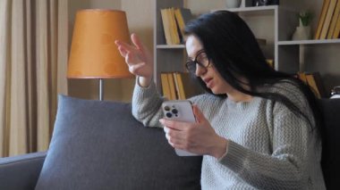 Genç, güzel bir kadın, evinde kanepede oturuyor, telefonda konuşuyor, duygusal olarak el kol hareketi yapıyor. Sıcak ev ortamı.