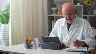 60 yaşındaki yaşlı bir erkek doktor bir masada oturur, bir bilgisayar üzerinde çalışır, tıbbi geçmişleri inceler, test sonuçlarını analiz eder, hastalara telefon eder, hasta için bir tedavi planı düşünür, reçete yazar..