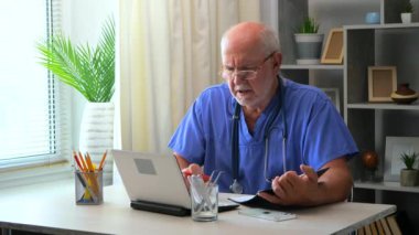 60 yaşındaki yaşlı bir erkek doktor bir masada oturur, bir bilgisayar üzerinde çalışır, tıbbi geçmişleri inceler, test sonuçlarını analiz eder, hastalara telefon eder, hasta için bir tedavi planı düşünür, reçete yazar..