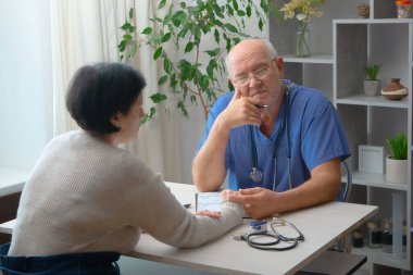 Yaşlı bir doktor, iş yerinde, hastalarını kabul eder. Engelli bir kadını muayene ediyor, parmak kemikleri kayıp..