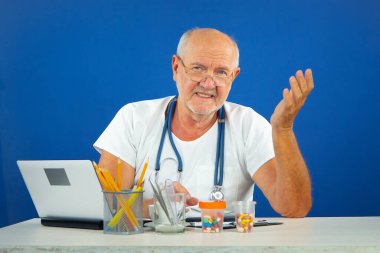 Yaşlı bir adam, bir doktor, ofisinde, masada. Mavi arkaplan.