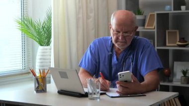 Yaşlı bir cerrah doktor bir ofiste bilgisayar ve akıllı telefonla çalışır, bir tedavi programı yapar, iyileşme sürecinin devamı için reçete yazar..