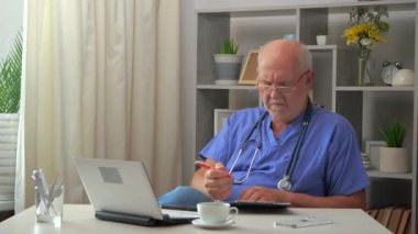 Yaşlı bir cerrah doktor hastanın tıbbi geçmişi olan bir ofiste çalışır, doktor tabletine veri kaydeder, tedaviyi ayarlar. Yavaş çekim.