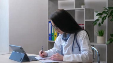 Genç, güzel bir kadın doktor bilgisayarında çalışıyor, hastanın sağlık bilgilerini kontrol ediyor ve veritabanına kaydediyor, ve düzeltmeleri bir deftere yazıyor..