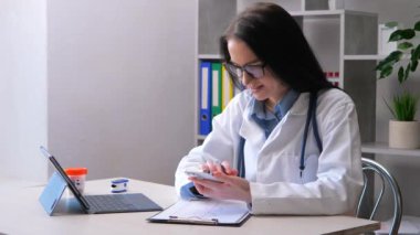 Genç, güzel bir doktor bilgisayar başında bir ofiste çalışıyor ve akıllı telefonuyla hastanın sağlığıyla ilgili verileri kontrol ediyor ve veri tabanına giriyor..