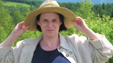 Orta yaşlı bir çiftçi kadın, güneşli bir yaz gününde açık havada, bir şapka takıyor, duygusal olarak gülümsüyor, yüksek dağların arka planına karşı. Yavaş çekim.