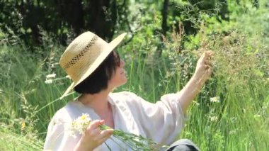 Beyaz bluzlu ve şapkalı orta yaşlı bir kadın çiftçi uzun çimlerde oturur, güneşli bir yaz gününün tadını çıkarır, açık havada. Yavaş çekim.