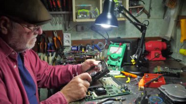 Eski bir usta atölyede bilgisayar parçaları tamir eder. Lehim demiri ve diğer aletleri kullanarak bozuk elektronikleri tamir eder ve ayarlar..
