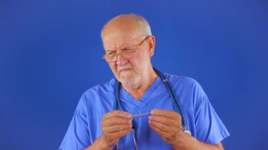 60 yaşında yaşlı bir doktor, mavi arka planda, hastanın vücut sıcaklığının termometre değerlerini kontrol ediyor. Yavaş çekim.