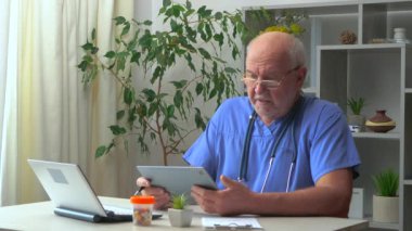 60 yaşındaki yaşlı bir doktor masaya oturur, bilgisayar üzerinde çalışır, tıbbi geçmişleri inceler, test sonuçlarını analiz eder, hastaları arar, tedavi planını düşünür, ilaç yazar, tabletle iletişim kurar..