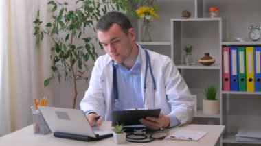 Genç bir stajyer doktor bir ofiste çalışıyor, tıbbi geçmişi inceliyor, teşhis koymak için bilgisayar kullanıyor ve uygun tedaviyi yazıyor. Yavaş çekim.