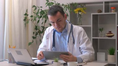 Genç bir stajyer doktor bir ofiste çalışıyor, bilgisayarlı tablet kullanarak hastanın tıbbi geçmişini araştırıyor, teşhisi düşünüyor ve uygun tedaviyi yazıyor. Yavaş çekim.