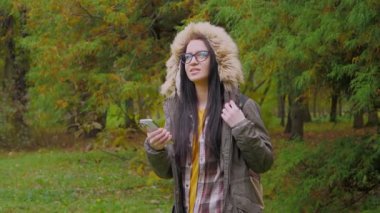 Doğada seyahat. Sonbahar ormanındaki genç güzel kız sonbahar doğasının güzelliğinin tadını çıkarıyor, yürüyüş yapıyor, akıllı telefondan arkadaşlarıyla iletişim kuruyor, gülümsüyor. Yavaş çekim.