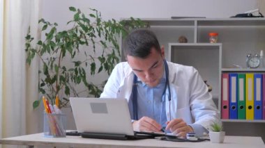 Genç bir stajyer doktor bir ofiste çalışıyor, hastanın tıbbi geçmişini, tedavi yöntemlerini ve tanısını bir bilgisayardan okuyor. Yavaş çekim.