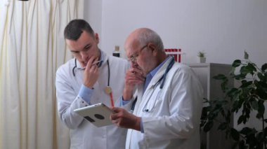 Beyaz önlüklü genç bir doktor yaşlı bir doktorla iletişim kuruyor, tedavi ilkelerini tartışıyor, ilaç yazıyor ve konsültasyon yapıyor. Yavaş çekim.