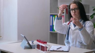 Genç bir kadın, bir hemşire, bir laboratuvar teknisyeni, analiz için alınan kanı inceler, test tüplerini numunelerle inceler, onları değerlendirir. Yavaş çekim.