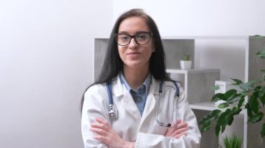 Genç bir kadın, doktor, steteskoplu ve laboratuvar önlüklü, kameraya gülümseyen, kollarını göğsüne kavuşturmuş. Yavaş çekim.