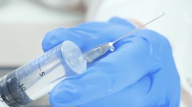 Mavi eldivenli doktor enjeksiyondan önce şırıngayı kontrol ederken iğneden sıkılan kan damlalar düşüyor. Eller yakın çekim. Yavaş çekim.