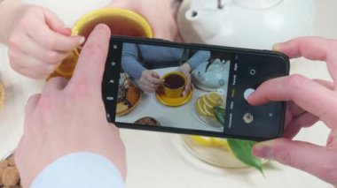 Genç bir kız, sıcak çayı kaşıkla karıştırıyor, mutfak masasında tatlı ve meyvelerle birlikte. Bir adamın elleri, akıllı telefonu tutuyor ve fotoğraf çekiyor. Kahvaltıda. Yakın plan. Yavaş çekim.
