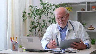 Yaşlı bir erkek doktor, doktorluk yapıyor, belgelerle uğraşıyor, hastalarla iletişim kuruyor, teşhis koyuyor, tedavi uyguluyor. Yavaş çekim.