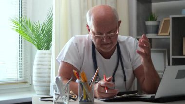 60 yaşındaki yaşlı bir doktor masaya oturur, bilgisayar üzerinde çalışır, tıbbi geçmişleri inceler, test sonuçlarını analiz eder, hastaları arar, tedavi planını düşünür, ilaç yazar, tabletle iletişim kurar..