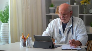 60 yaşındaki yaşlı bir doktor masaya oturur, bilgisayar üzerinde çalışır, tıbbi geçmişleri inceler, test sonuçlarını analiz eder, hastaları arar, tedavi planını düşünür, ilaç yazar, tabletle iletişim kurar..