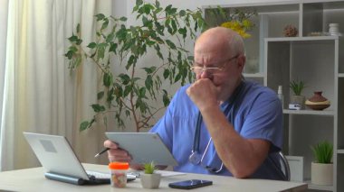 60 yaşındaki yaşlı bir doktor masaya oturur, bilgisayar üzerinde çalışır, tıbbi geçmişleri inceler, test sonuçlarını analiz eder, hastaları arar, tedavi planını düşünür, ilaç yazar, tabletle iletişim kurar..