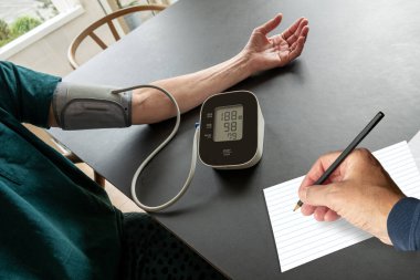 Kişi, kan basıncını ev ortamında bir masada bir başka kişinin bir kağıt parçasına not tutmasını izlerken manşet ve elektronik monitörle ölçüyor. Yüksek tansiyon sağlık konsepti.