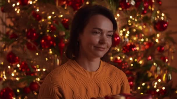 バックグランドクリスマスツリー上で幸せな笑顔ギフトと赤いカートンを保持白人女性装飾されたライトが点滅し 輝く魔法の新しい年前夜 — ストック動画