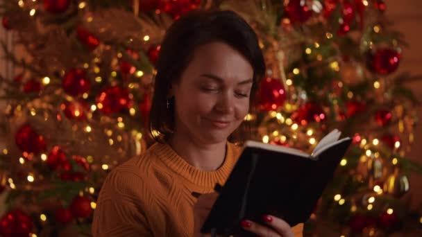 快乐的女人拿着笔在笔记本上写字 新年决心或在快乐杂志上写笔记 写在背景圣诞树上 灯闪闪着 闪着光芒 — 图库视频影像