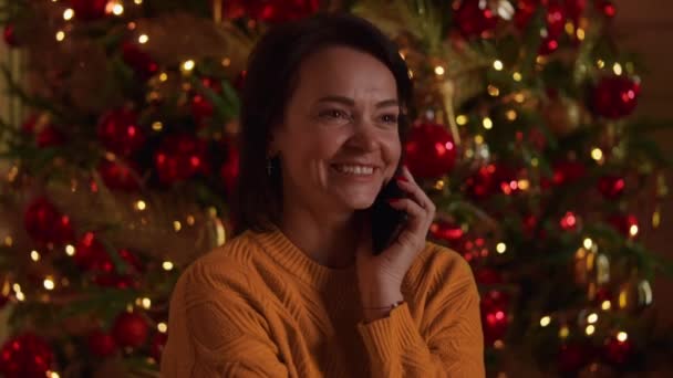 微笑的女人拿着智能手机和朋友聊天 身穿针织毛衣 圣诞树装饰的灯火通明 迷人的魔法新年前夕 — 图库视频影像