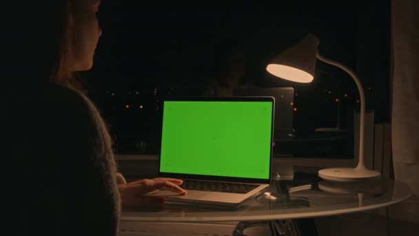 在晚上上网或在公寓里上网购物时 使用绿色屏幕笔记本电脑的女人坐在大橱窗前的书桌前 — 图库视频影像