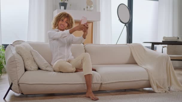 スマートフォンでアプリを使用してアパートのソファーに座って自宅でセルフィー携帯電話を取る若い女性 — ストック動画
