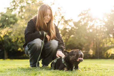 Genç bir kız çimlerin üzerinde Fransız bulldog köpeğine sarılıyor..