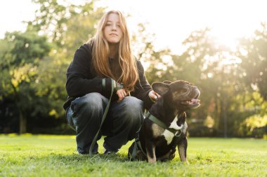 Genç bir kız çimlerin üzerinde Fransız bulldog köpeğine sarılıyor..