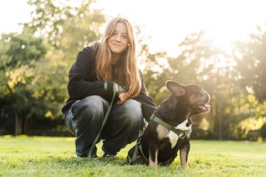 Genç bir kız çimlerin üzerinde Fransız bulldog köpeğine sarılıyor..