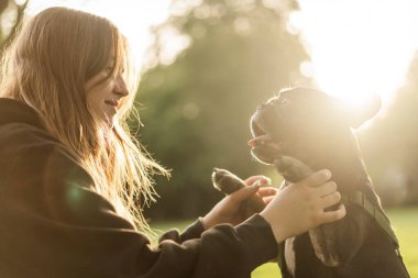 Genç bir kız çimlerin üzerinde Fransız bulldog köpeğine sarılıyor..