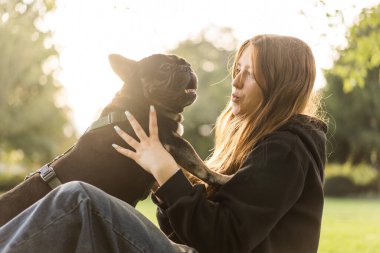 Genç bir kız çimlerin üzerinde Fransız bulldog köpeğine sarılıyor..