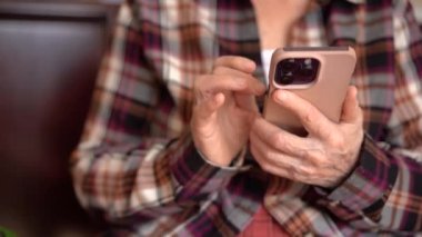 Seksen yaşında bir kadın elinde telefon, evde çalışıyor. Telefonla ellerin yakın plan görüntüsü
