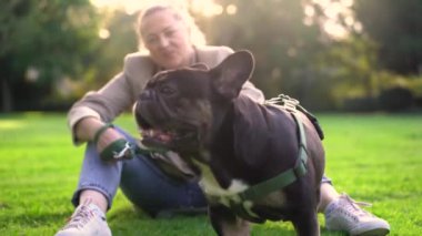 40 'lı yaşlarda bir kadın, köpeği Fransız bulldog' la parkta çimlerin üzerinde oynuyor. 4K görüntülerinde.