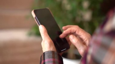 Seksen yaşında bir kadın elinde telefon, evde çalışıyor. Telefonla ellerin yakın plan görüntüsü