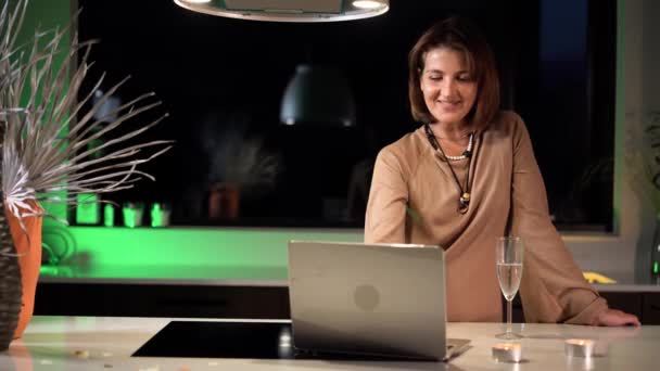 キッチンの自宅の女性フリーランサーは 緑の照明4Kビデオの背景に対して お祝いのドレスでラップトップでロマンチックな会話をしています — ストック動画