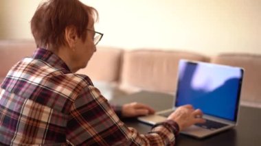 Yaşlı kadın 80 yaşında dizüstü bilgisayarıyla evde çalışıyor. Mavi dizüstü bilgisayar ekranı.