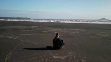 Siyah elbiseli ve dizüstü bilgisayarlı bir kız İzlanda 'da siyah bir sahilde oturuyor. Özgürlük ve serbest dolaşım kavramı, İHA 'nın 4K görüntüsü.