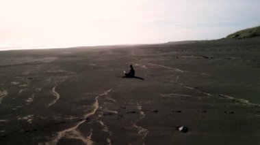 Siyah elbiseli ve dizüstü bilgisayarlı bir kız İzlanda 'da siyah bir sahilde oturuyor. Özgürlük ve serbest dolaşım kavramı, İHA 'nın 4K görüntüsü.