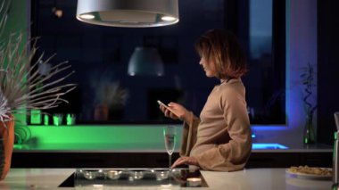 Mutfakta telefonu olan bir kadın, yeşil ışıklandırma videosunun arka planına karşı, neşeli bir elbise içinde telefon ve romantik bir sohbet bekliyor.